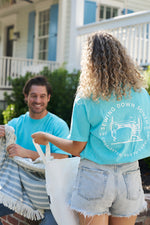 Two people outside a house in matching turquoise Sewing Down South Logo Rainbow Row Tees, one sitting with striped fabric and the other standing with a tote bag. The porch and lush greenery evoke the charm of Rainbow Row in the backdrop.
