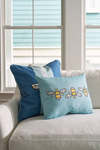 Two decorative pillows rest on a white sofa. The front pillow is a Bee Trio Lumbar Pillow from the Bee & Florals Collection, featuring a light blue surface with yellow bees. The back pillow is dark blue with light blue stripes. A window reveals part of a turquoise house.