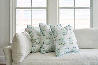 A chic light gray sofa is adorned with three Bee & Florals Collection: Victorian Floral in Blue Pillows, showcasing a green and blue Victorian floral pattern. Positioned in front of large windows, it bathes the room in natural light and is from Sewing Down South.