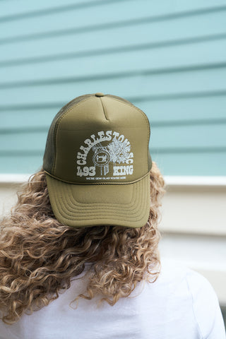 A person with curly blonde hair wears the Olive King St Trucker, an adjustable snapback in olive green with white CHARLESTONS 493 KING text and a small graphic, standing before a light blue wall resembling a Charleston storefront.