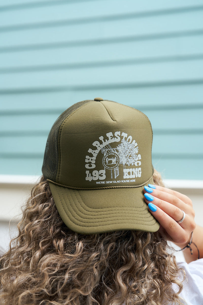 A person with curly hair wears an Olive King St Trucker hat with white CHARLESTOWNE KING 493 JI and WERE SEW GLAD YOURE HERE text, posed in front of a light blue Charleston storefront-inspired wall.