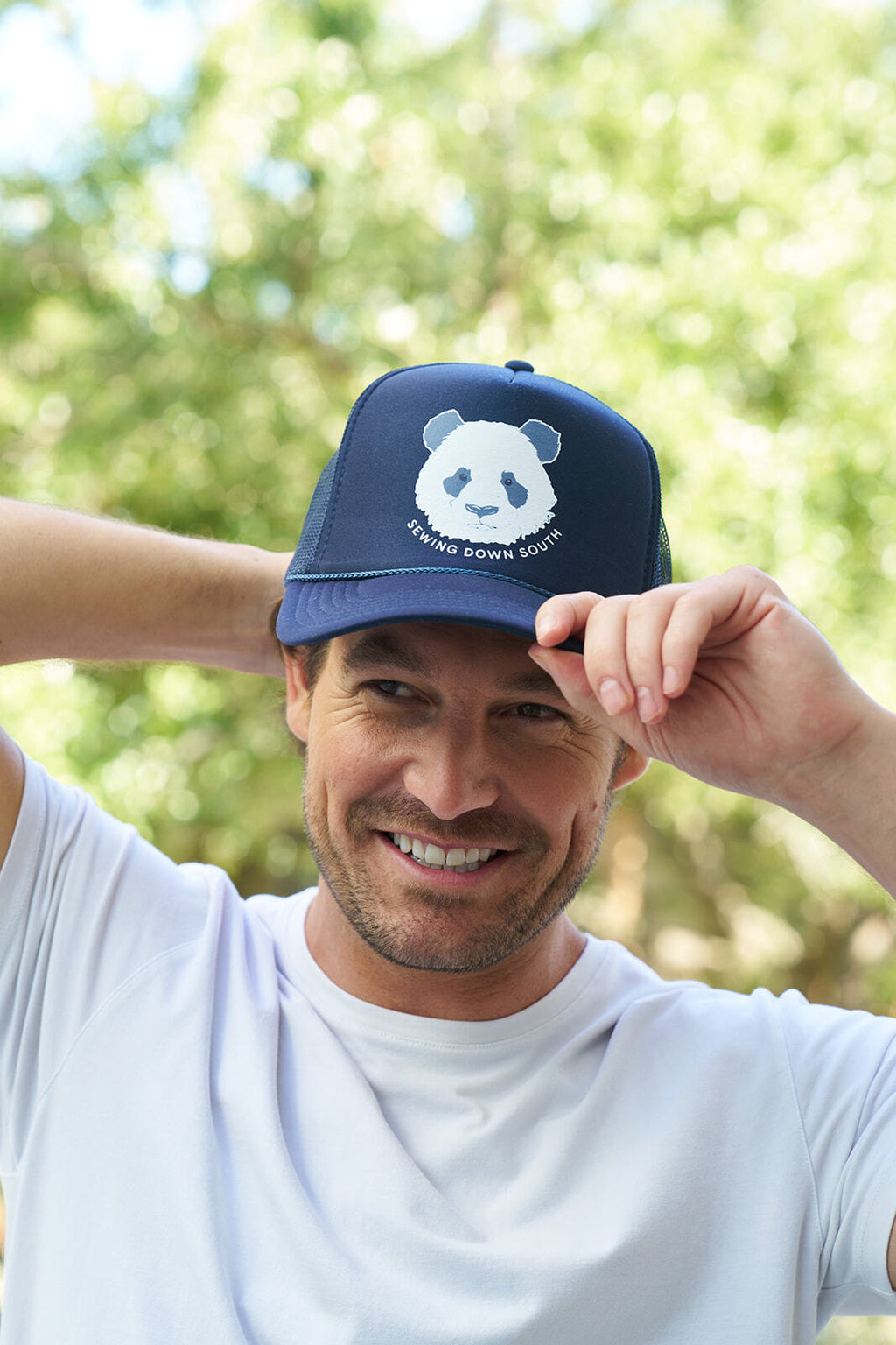 A smiling man stands outdoors in a white t-shirt and a navy blue Panda Trucker Hat with an adjustable snapback, featuring a panda face logo and SPINNING DOWN SOUTH text, with greenery in the background.