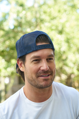 A man with a short beard and mustache wears the Panda Trucker Hat and a white t-shirt, smiling outdoors with green trees softly blurred in the background.