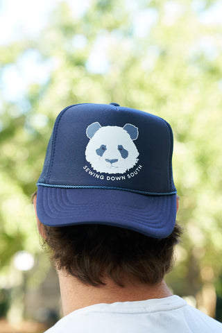 A person wears the Panda Trucker Hat, a navy blue adjustable snapback with a white panda face and Sewing Down South below it, perfect for Panda People. The background features blurred greenery outdoors.