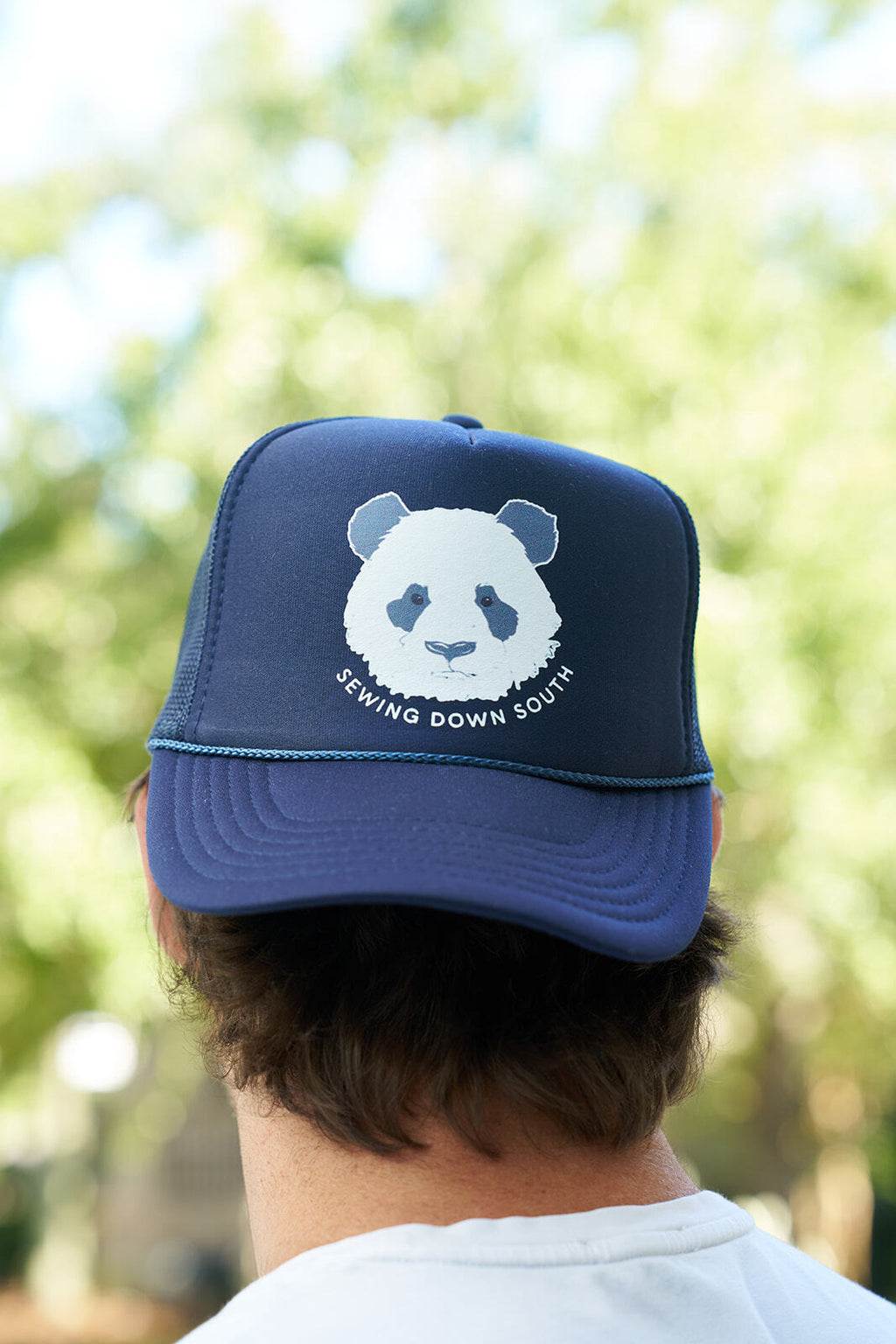 A person wears the Panda Trucker Hat, a navy blue adjustable snapback with a white panda face and Sewing Down South below it, perfect for Panda People. The background features blurred greenery outdoors.