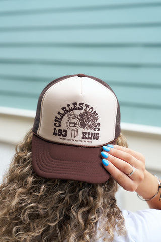 A person with curly hair wears the SDS King Street Trucker Hat in brown and white. Holding the brim with one hand, they display blue-painted nails against a pastel blue backdrop.