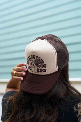 With long hair and pink nails, a person adjusts the SDS King Street Trucker Hat from the Charleston Storefront collection, labeled Charles Towne Landing 1670 with an old sailing ship graphic, against a light blue paneled wall.