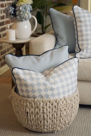 An Oversized Mod Dot Grid Pillow in light blue and white with hand-drawn patterns sits in a chunky knitted beige basket by a cream sofa, beside a side table with a pitcher and flowers—a modern Southern decor accent.