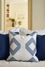 A close-up of the Mod Geo Diamond in Navy Pillow, featuring a navy and white geometric design, sits between solid navy pillows on a beige sofa; blurred plants and framed art in the background add Southern charm.