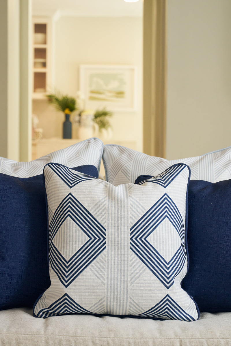 A close-up of the Mod Geo Diamond in Navy Pillow, featuring a navy and white geometric design, sits between solid navy pillows on a beige sofa; blurred plants and framed art in the background add Southern charm.