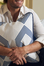 A man in a white shirt sits and smiles, holding the Mod Geo Diamond in Navy Pillow with its chic blue and white geometric pattern. Only his lower face and arms are visible, showcasing this modern accent ideal for contemporary decor.