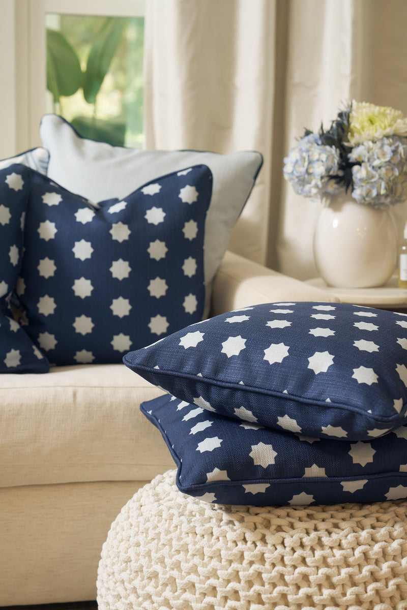 A Mod Stars Pillow, featuring navy blue fabric with white star patterns, rests on a beige sofa and knitted pouf. A vase of flowers sits nearby as sunlight streams through the window in the background.