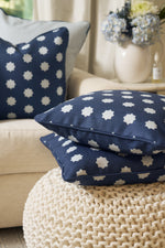 Two navy blue Mod Stars Pillows with white star designs are on a chunky knit ottoman, while a matching blue accent pillow decorates the beige sofa behind them. A vase of flowers is placed on a nearby table.