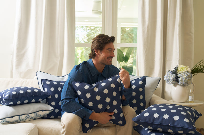 A man smiles on a light sofa, holding the Mod Stars Pillow—a blue accent pillow with white star patterns—surrounded by geometric pillows. Nearby, a vase of flowers sits on a table, with large windows in the background.