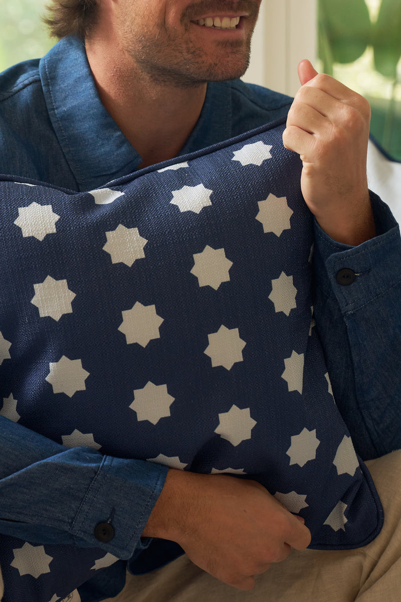 A man in a blue button-up shirt smiles while hugging the Mod Stars Pillow, which showcases a navy-blue and white geometric star pattern.