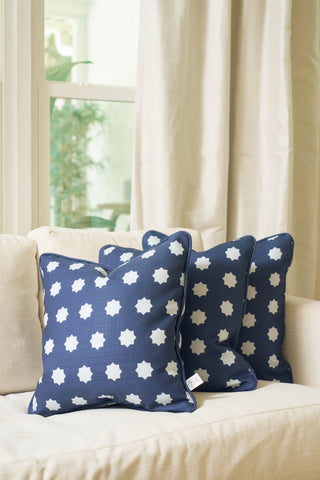 Three Mod Stars Pillows in navy blue with white star patterns are arranged on a light beige sofa by a window with cream curtains, as natural light brightens the cozy, modern living space.