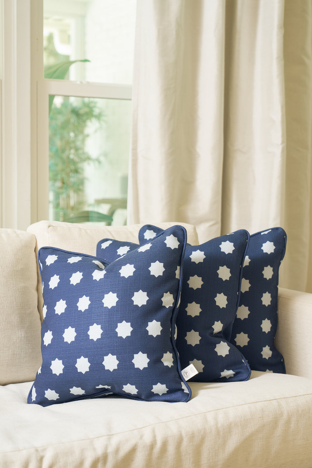 Three Mod Stars Pillows in navy blue with white star patterns are arranged on a light beige sofa by a window with cream curtains, as natural light brightens the cozy, modern living space.
