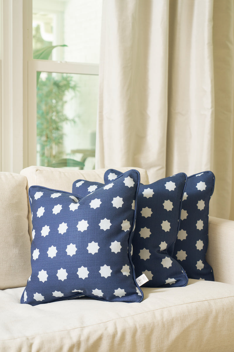 Three Mod Stars Pillows in navy blue with white star patterns are arranged on a light beige sofa by a window with cream curtains, as natural light brightens the cozy, modern living space.