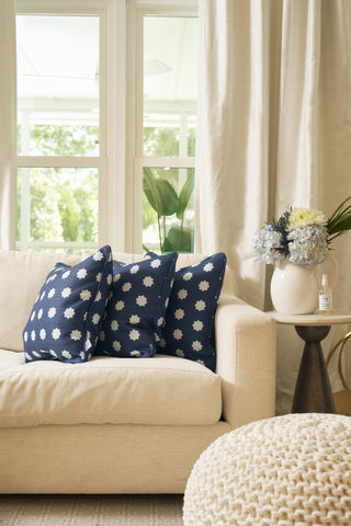 A cream sofa with two navy blue pillows featuring white floral patterns and a Mod Stars Pillow sits by a round side table holding a white pitcher with hydrangeas and a small bottle, in a bright room with large windows and curtains.