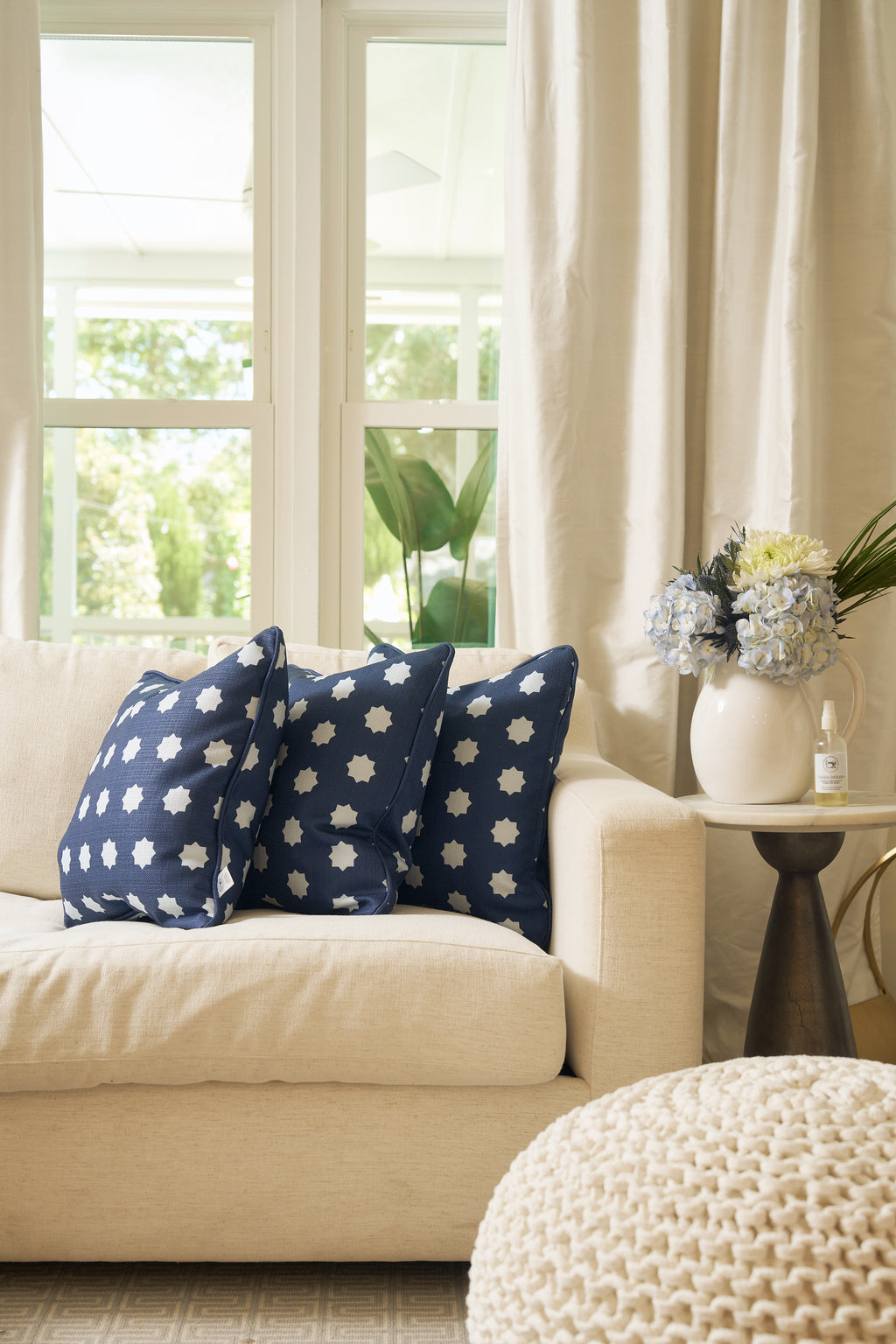 A cream sofa with two navy blue pillows featuring white floral patterns and a Mod Stars Pillow sits by a round side table holding a white pitcher with hydrangeas and a small bottle, in a bright room with large windows and curtains.