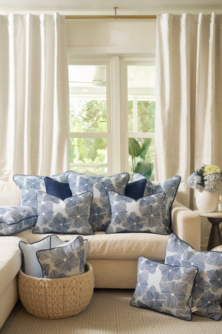 A light sofa features several blue and white floral-patterned pillows, including the Mod Flora Lumbar Pillow, in front of large windows with white curtains. A woven basket and hydrangeas finish the bright, airy Southern decor.