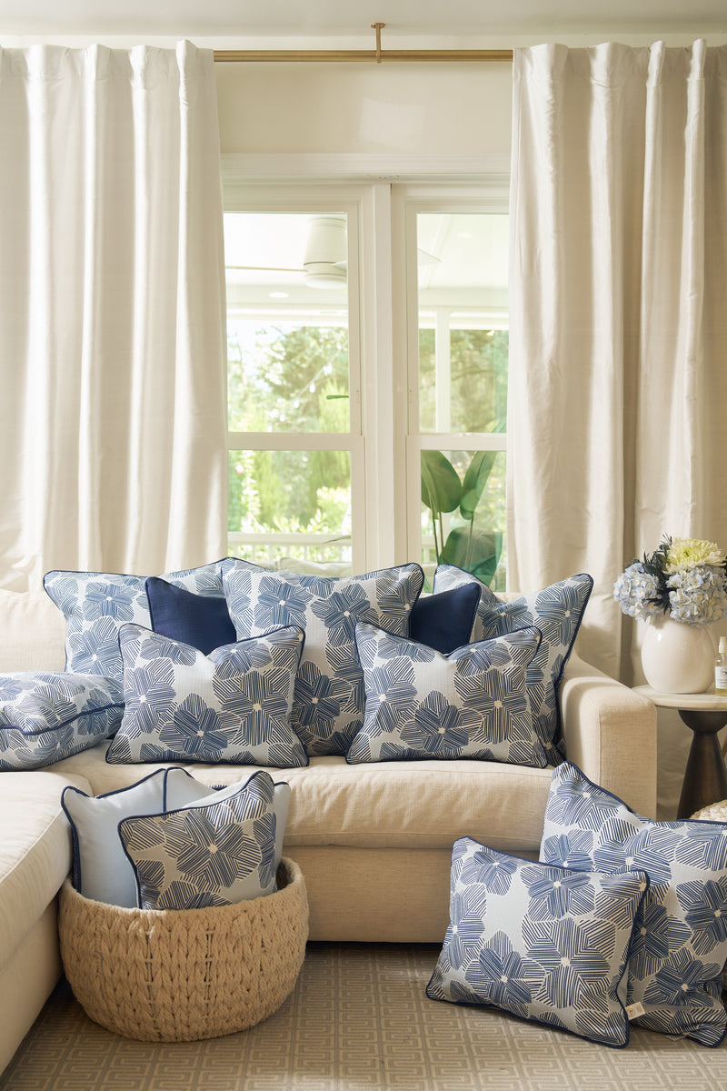 A light sofa features several blue and white floral-patterned pillows, including the Mod Flora Lumbar Pillow, in front of large windows with white curtains. A woven basket and hydrangeas finish the bright, airy Southern decor.