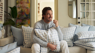 A man with brown hair and a beard, wearing a light gray sweater, sits on a couch holding a patterned pillow. The couch is covered with similar light blue and white pillows. The room is decorated with modern art and plants.