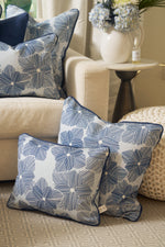 Two Mod Flora Lumbar Pillows featuring blue and white geometric floral patterns rest on a beige carpet beside a cream sofa, with another matching pillow and a floral vase in the background, highlighting Southern charm.