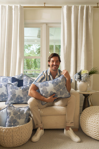 A smiling man sits on a beige sofa holding a Mod Flora Lumbar Pillow, surrounded by matching modern pillows. White curtains, a window, and a side table with flowers and a plant complete the cozy Southern-inspired decor.