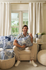 A smiling man sits on a beige sofa holding a Mod Flora Lumbar Pillow, surrounded by matching modern pillows. White curtains, a window, and a side table with flowers and a plant complete the cozy Southern-inspired decor.