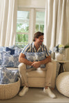 A man in a blue-and-white striped shirt sits on a cream sofa, hugging the Mod Flora Lumbar Pillow. Blue and white cushions and floral accents complete the Southern-style living room with large windows and white curtains.