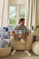 A man in a blue-and-white striped shirt sits on a cream sofa, hugging the Mod Flora Lumbar Pillow. Blue and white cushions and floral accents complete the Southern-style living room with large windows and white curtains.
