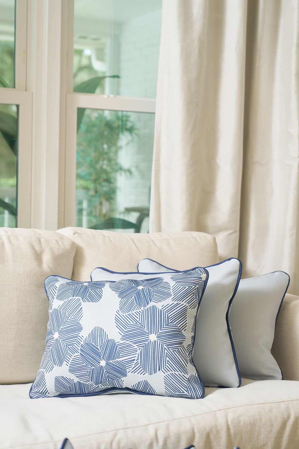 A beige sofa with Southern decor features two pillows—one is the Mod Flora Lumbar Pillow, known for its blue and white geometric floral design, while the other is a solid light blue modern pillow—arranged before tall windows with cream curtains.