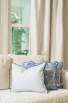 A beige sofa with a Mod Flora Lumbar Pillow and two blue and white patterned pillows sits before a window framed by cream curtains and greenery, creating inviting Southern decor.