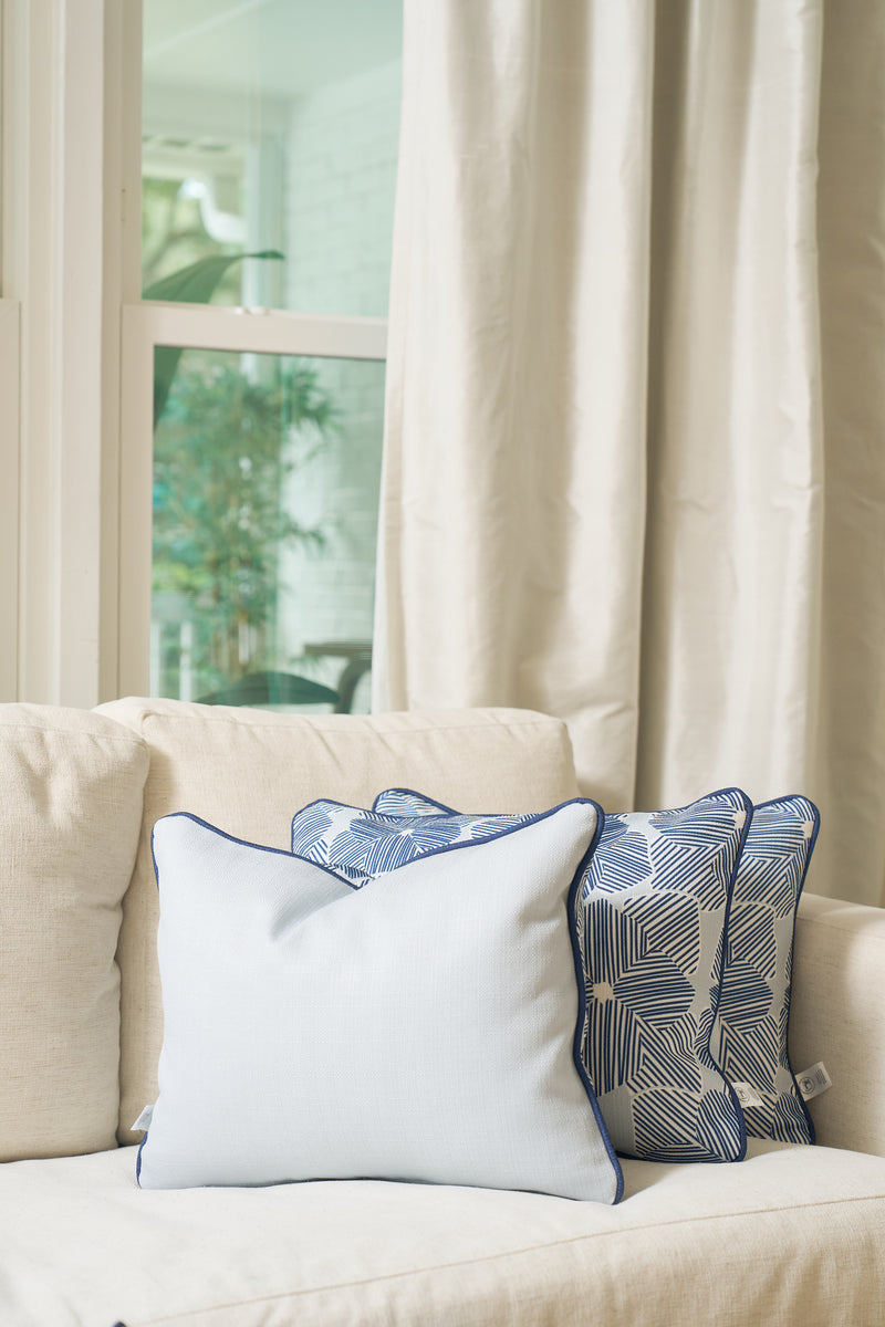 A beige sofa with a Mod Flora Lumbar Pillow and two blue and white patterned pillows sits before a window framed by cream curtains and greenery, creating inviting Southern decor.