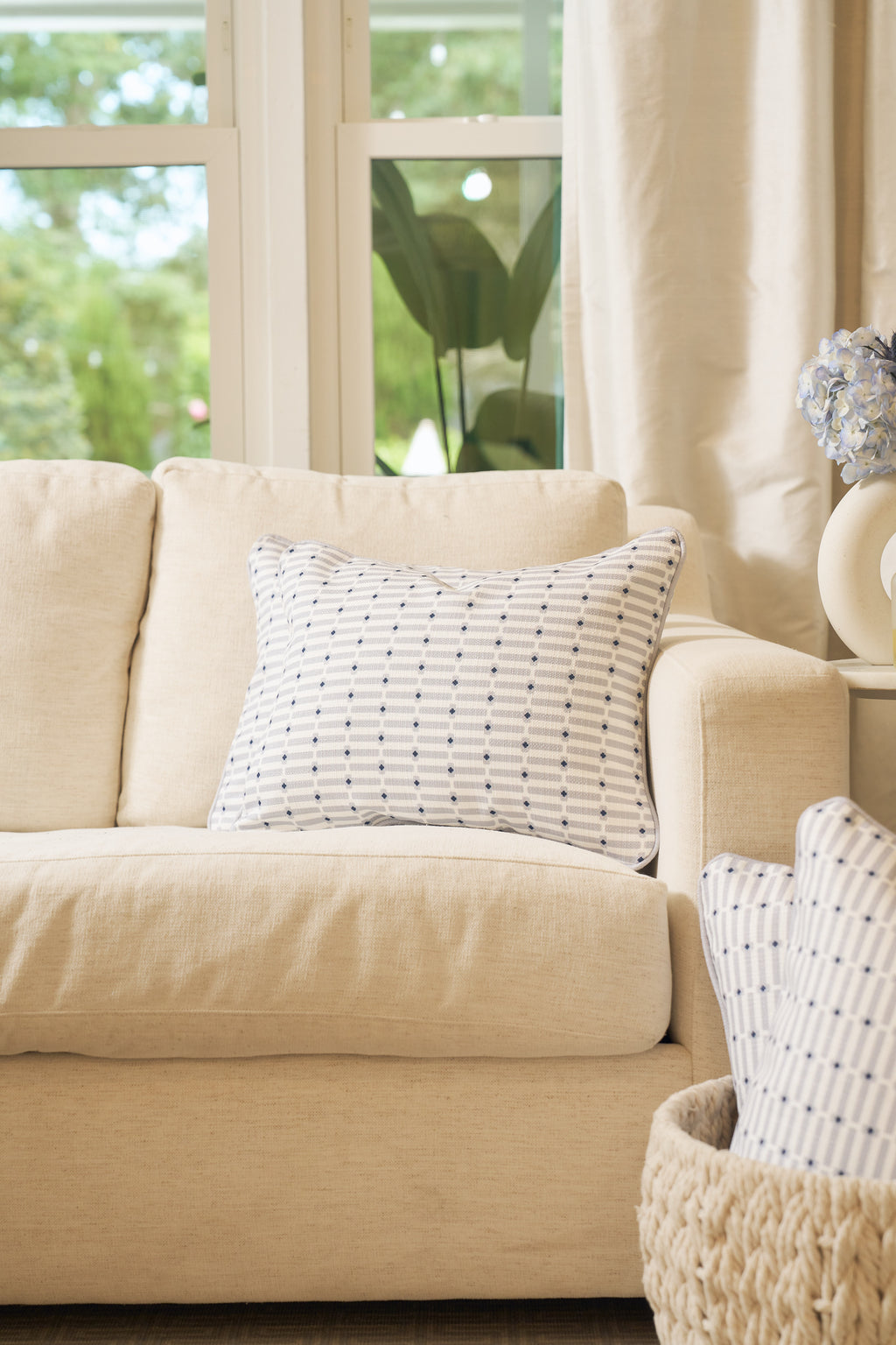 A beige sofa with the Mod Mini Crossing Tiles Lumbar Pillow sits by a sunlit window. A woven basket holds an accent pillow in front, while flowers and neutral curtains complete this Southern-inspired space.