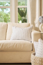 A beige sofa with the Mod Mini Crossing Tiles Lumbar Pillow sits by a sunlit window. A woven basket holds an accent pillow in front, while flowers and neutral curtains complete this Southern-inspired space.
