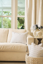 A cozy living room features a beige sofa with the Mod Mini Crossing Tiles Lumbar Pillow, a round side table topped with flowers, a woven basket holding an extra pillow, and large windows draped in cream curtains for natural light.