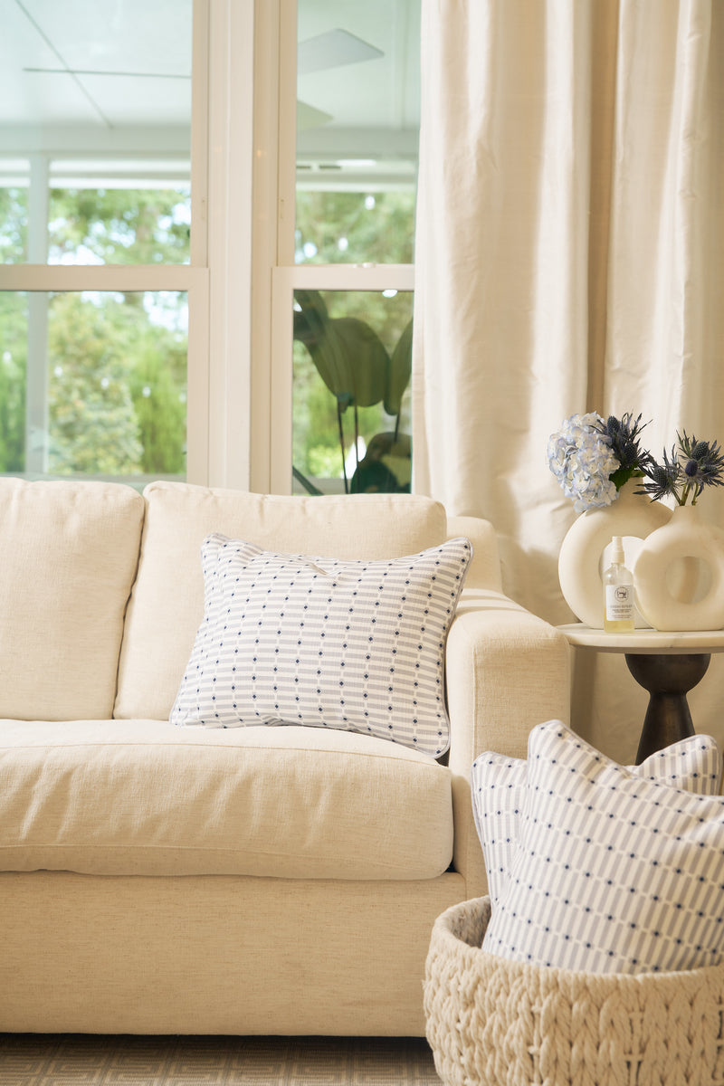 A cozy living room features a beige sofa with the Mod Mini Crossing Tiles Lumbar Pillow, a round side table topped with flowers, a woven basket holding an extra pillow, and large windows draped in cream curtains for natural light.
