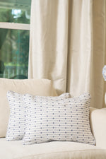 Two Mod Mini Crossing Tiles Lumbar Pillows, featuring a blue dotted grid pattern, rest on a cream sofa by a window with sheer curtains. Sunlight fills the cozy space and adds a touch of Southern decor charm.