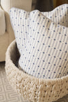 Two Mod Mini Crossing Tiles Lumbar Pillows with a blue and white checkered pattern rest in a chunky knit beige basket beside a light-colored upholstered chair, adding a Southern decor touch.
