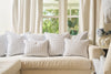 A beige sectional sofa topped with white and blue pillows, featuring the Mod Mini Crossing Tiles Lumbar Pillow, sits before large windows with light curtains, creating a cozy living room filled with natural light and Southern charm.