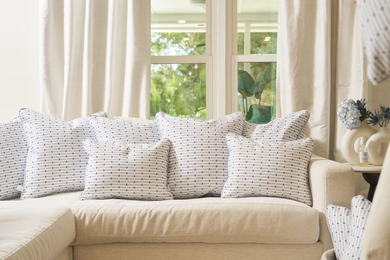 A beige sectional sofa topped with white and blue pillows, featuring the Mod Mini Crossing Tiles Lumbar Pillow, sits before large windows with light curtains, creating a cozy living room filled with natural light and Southern charm.