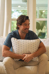 A man with wavy brown hair and a beard smiles on a couch, hugging the Mod Mini Crossing Tiles Lumbar Pillow. He wears a blue short-sleeve shirt and beige pants, with sunlight streaming through large windows in a relaxed Southern setting.