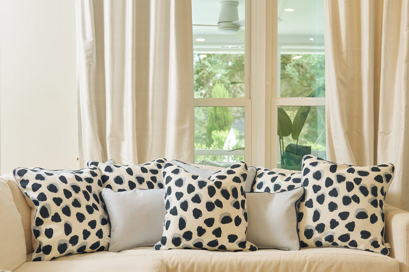 A beige sofa with five decorative pillows, including three Oversized Modern Polka Dots Pillows and two plain light-colored ones, sits by large windows with cream curtains. Greenery outside brings a fresh feel to this inviting southern decor.