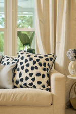 A beige sofa with Oversized Modern Polka Dots Pillows in white and navy sits by a window with cream curtains. A side table holds a blue hydrangea bouquet and bottle, adding southern charm while greenery peeks in from outside.