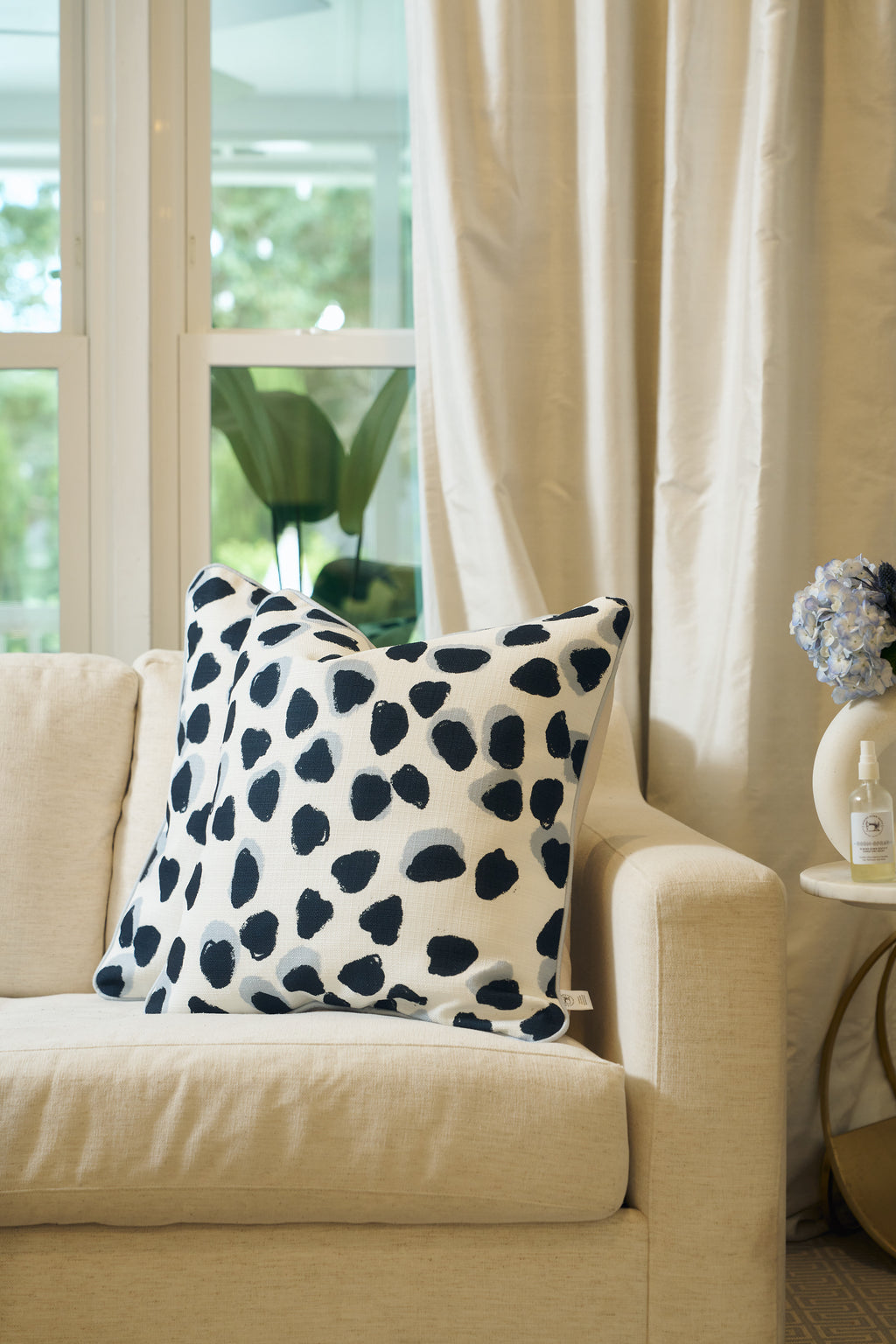 A cream-colored sofa with an Oversized Modern Polka Dots Pillow sits by a round side table with blue hydrangeas and lotion in a bright, window-lit room decorated with light curtains.