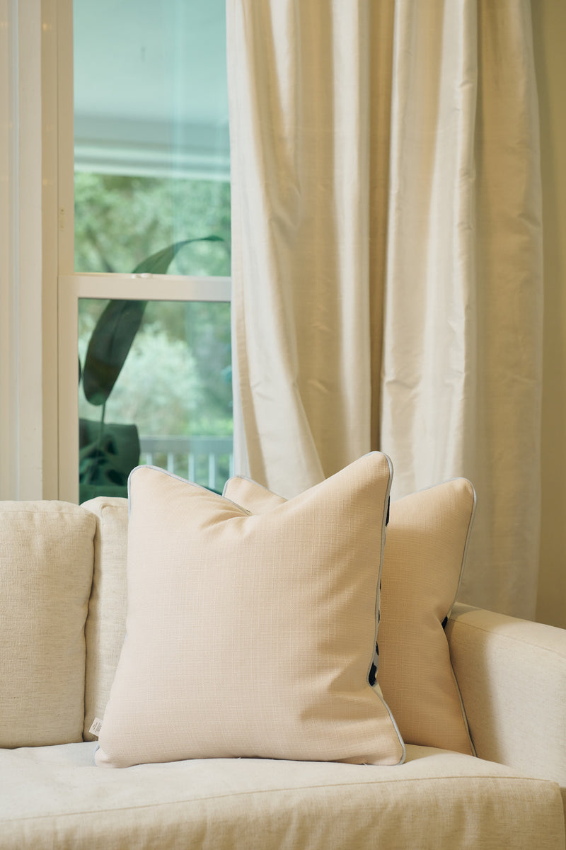 A beige sofa features an Oversized Modern Polka Dots Pillow and a light cream pillow, set before a window with white curtains. Sunlight streams in, illuminating the neutral-toned living space with greenery visible outside.