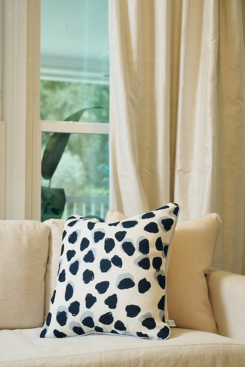 A cream-colored sofa with the Oversized Modern Polka Dots Pillow, featuring a hand-painted pattern, sits by a window with sheer beige curtains and views of lush greenery, adding a hint of Southern decor.
