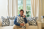 A man sits on a beige sofa, smiling and holding an Oversized Modern Polka Dots Pillow with a hand-painted pattern. Similar black-spotted pillows line the sofa, enhancing the southern decor. White curtains frame a window overlooking greenery.